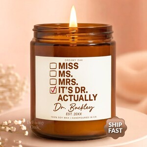 May include: A lit brown glass jar candle with a white label that reads "IT'S DR. ACTUALLY" with the name "Dr. Buckley" below. The candle is on a pink surface with a soft pink background.