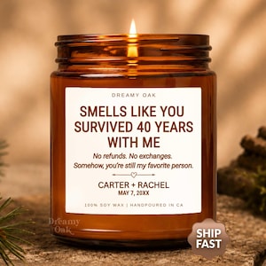 May include: A lit candle in an amber glass jar with a white label that reads: DREAMY OAK, SMELLS LIKE YOU SURVIVED 40 YEARS WITH ME, No refunds. No exchanges. Somehow, you're still my favorite person. CARTER + RACHEL, MAY 7, 20XX, 100% SOY WAX.