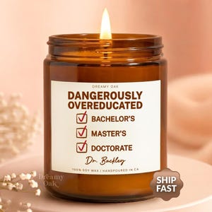 May include: A lit brown glass candle jar with a white label that reads "DANGEROUSLY OVEREDUCATED" with checkmarks next to "Bachelor's", "Master's", and "Doctorate". The candle is labeled "Dreamy Oak" and is hand-poured in CA.