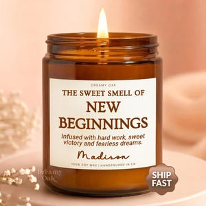 May include: A lit brown glass jar candle with a white label. The label reads "THE SWEET SMELL OF NEW BEGINNINGS" and "Infused with hard work, sweet victory and fearless dreams." The candle is labeled "Madison" and "100% SOY WAX HANDPOURED IN CA."