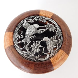 May include: A round wooden bowl with a decorative metal insert featuring two hummingbirds and floral accents. The bowl has a dark brown and light brown wood grain pattern. The metal insert is silver-colored.