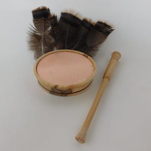 May include: A wooden turkey call with a light brown finish, featuring a circular sound chamber with a pink interior, a wooden striker, and a cluster of brown and black feathers. The call is designed for hunting.