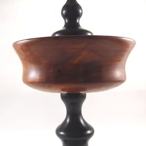 May include: A wooden bowl with a dark brown finish sits on a black wooden pedestal with a pointed finial. The bowl is wide and shallow, with a slightly flared rim.