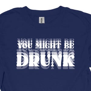 以下が含まれることがあります： ネイビーブルーのTシャツ。白い文字で「YOU MIGHT BE DRUNK」と縦に重ねて表示され、影の効果があります。カジュアルな服装アイテムです。