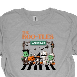 以下が含まれることがあります： モンスターが横断歩道を歩いている漫画のグラフィックが描かれたグレーのTシャツ。モンスターの上の看板には「The Boo-Tles」と「Scabby Road」の文字が書かれています。
