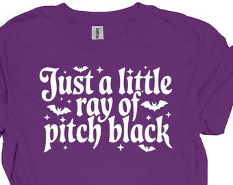 May include: A purple t-shirt featuring the text "Just a little ray of pitch black" in white, with bat illustrations. The tee is a solid purple colour and has short sleeves.