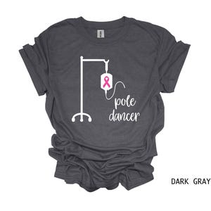 May include: Dark gray t-shirt with a white graphic of an IV pole and the text "pole dancer". The IV bag has a pink ribbon.