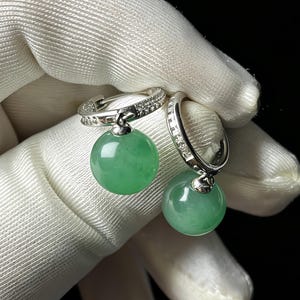 Peut inclure: Une paire de boucles d'oreilles créoles en argent avec une rangée de petites pierres transparentes et une pierre de jade verte claire ronde et pendante. Les boucles d'oreilles sont présentées sur fond blanc.