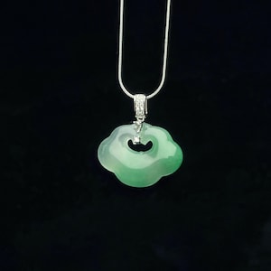 Puede incluir: Collar con colgante de jade con una piedra de jade en forma de nube. El jade es de color verde claro, con una cadena de plata y una pequeña fianza con diamantes. El colgante cuelga sobre un fondo negro.
