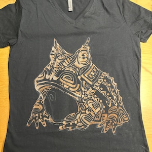 Könnte beinhalten: Schwarzes T-Shirt mit V-Ausschnitt und einem braunen, tribalen Frosch-Design.