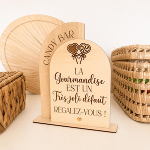 Könnte beinhalten: Ein helles, bogenförmiges Holzschild mit flachem Sockel, graviert mit dem Text CANDY BAR, LA GOURMANDISE EST UN TRÈS JOLI DÉFAUT RÉGALEZ-VOUS!, einem kleinen Herzsymbol und zwei Lutscher-Illustrationen. Das Schild hat eine glatte Holzoberfläche.