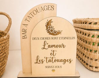 May include: A light-colored wooden sign with two arched panels. The taller panel reads BAR À TATOUAGES. The shorter panel features a crescent moon with flowers and the text DEUX CHOSES SONT ETERNELLES L'amour et Les tatouages SERVEZ-VOUS with a heart.