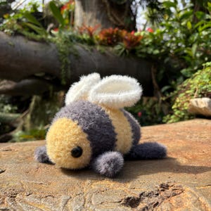 May include: A soft toy bee with a yellow head, grey body, and white wings. The bee has a black eye and is lying on a stone surface. The toy is made of a fuzzy material.