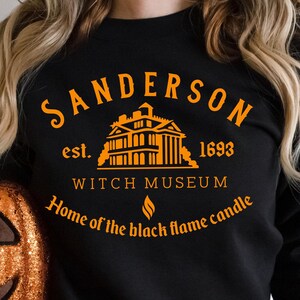 May include: Black sweatshirt with orange text that reads "Sanderson Witch Museum" and "Home of the black flame candle." An orange glitter pumpkin is held in front of the sweatshirt. The sweatshirt design includes a building graphic and the year 1693.
