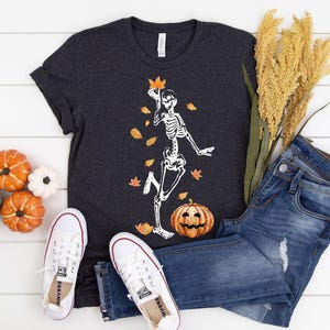 May include: A dark grey t-shirt featuring a white skeleton design, orange leaves, and a pumpkin. The outfit includes blue jeans and white trainers. Decorative pumpkins and wheat stalks complete the autumn theme.