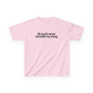 May include: A light pink short-sleeved t-shirt featuring the black text "AI could never recreate my swag" printed in a simple sans-serif font across the centre of the chest. The garment has a crew neckline and a standard straight hemline.