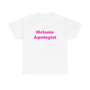 Könnte beinhalten: Ein weißes T-Shirt mit kurzen Ärmeln und dem Text Melanie Apologist, der in einer kräftigen, leuchtend rosa Schrift mittig auf der Brust aufgedruckt ist. Das Kleidungsstück hat einen klassischen Rundhalsausschnitt und einen geraden Saum.