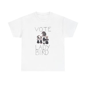 Op de afbeelding: Wit t-shirt met de zwarte tekst "VOTE LADY BIRD", met een afbeelding van een duif en verschillende figuren. De figuren dragen zwarte jurken en staan in een rij. Het t-shirt is gemaakt van een zacht materiaal.