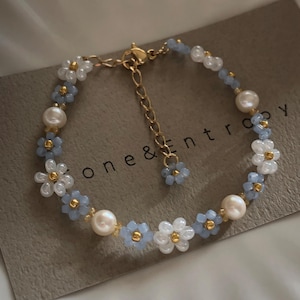 May include: A delicate bracelet featuring a floral design with white and light blue beads, interspersed with pearl accents. The bracelet has a gold-colored chain and clasp. The text "one & entropy" is visible on the background.