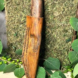 May include: A handcrafted knife with a wooden handle and a leather sheath. The handle is a dark brown color with a wood grain pattern. The sheath is a lighter brown with decorative stitching and carved details. The knife is displayed against a backdrop of green moss and leaves.