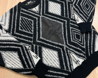 May include: Black and white geometric patterned sweater with a black leather patch on the front. The sweater has a crew neck and long sleeves.