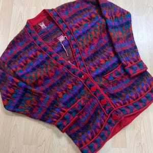 May include: A vintage cardigan in a vibrant geometric pattern of red, purple, blue, and orange. The sweater has a V-neck, red trim, and red buttons down the front. Two pockets are visible.