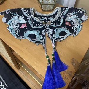 May include: A black embroidered collar with a dragon design and blue tassels. The collar is decorated with pearl beads and green accents.