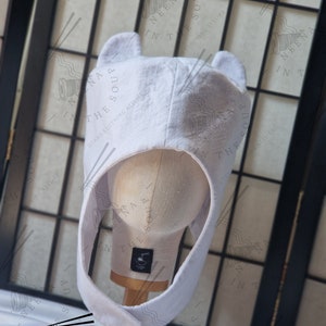 May include: White felt hat with two floppy ears, resembling a cartoon character. The hat is on a white mannequin head, with a wooden base. The hat is designed to look like a character from the show "Adventure Time".