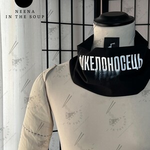 May include: A black neck gaiter with the white Cyrillic text "ИКЕЛОНОСЕЦЬ" is displayed on a mannequin. The mannequin is wearing a tan-colored long-sleeved shirt. The brand logo "NEENA IN THE SOUP" is visible.