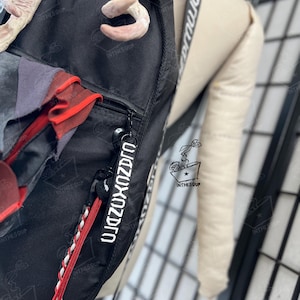 May include: A black backpack featuring a white strap with black text, a zip pull with white text, and a red and white braided cord. The bag is adorned with a light-coloured, textured antler-like attachment at the top and red fabric accents.