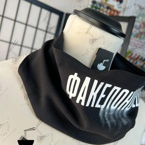 May include: Black neck gaiter with white Cyrillic text and a faded white design. The gaiter is displayed on a mannequin head. The brand name "NEENA IN THE SOUP" is visible on the tag and in the background.