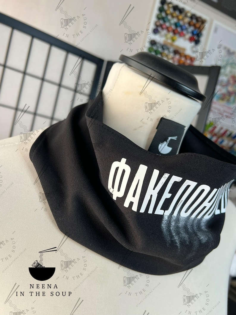 May include: Black neck gaiter with white Cyrillic text and a faded white design. The gaiter is displayed on a mannequin head. The brand name "NEENA IN THE SOUP" is visible on the tag and in the background.