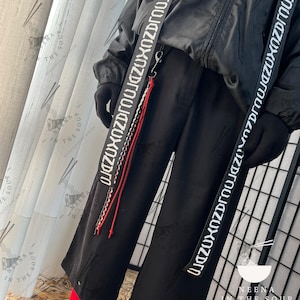 May include: Black outfit with wide-leg trousers and a black jacket. The outfit includes white straps with text and a red and white cord. The outfit is paired with black boots and red socks. The text on the straps reads "NEENA IN THE SOUP OTAKU CLOTHING ATELIER".