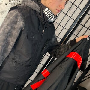 May include: Black hooded jacket with gray camouflage sleeves and a black scarf with white text. The jacket has a zipper closure and a pocket on the chest. A black garment with red bands is held in the hand.
