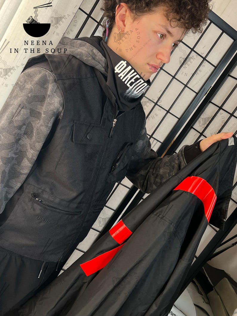 May include: Black hooded jacket with gray camouflage sleeves and a black scarf with white text. The jacket has a zipper closure and a pocket on the chest. A black garment with red bands is held in the hand.