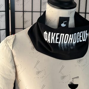 May include: A black neck gaiter with white Cyrillic text and a small tag. The text reads "ФАКЕЛОНОСЕЦЬ". The tag features a bowl of noodles with chopsticks. The gaiter is displayed on a mannequin.