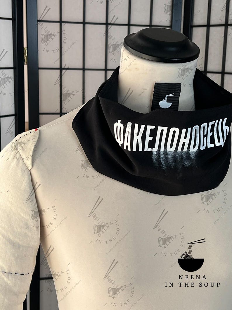 May include: A black neck gaiter with white Cyrillic text and a small tag. The text reads "ФАКЕЛОНОСЕЦЬ". The tag features a bowl of noodles with chopsticks. The gaiter is displayed on a mannequin.