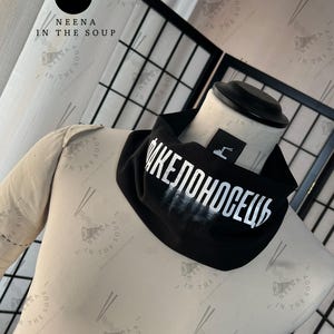 May include: Black neck gaiter with white Cyrillic text. The gaiter is displayed on a mannequin. The brand logo "NEENA IN THE SOUP" is visible in the background.