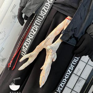 Può includere: Outfit nero con un elemento decorativo in legno chiaro. L'outfit include una giacca nera, pantaloni e una cinghia con testo bianco. Un logo con testo è visibile in basso.