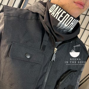 May include: Black vest with multiple pockets and a zipper closure. The vest has a button on the pocket and a small zipper pocket. A black neck gaiter with white text is visible. The text on the neck gaiter reads "FAKEПОНЧИ". The vest is part of the "Neena in the Soup" otaku clothing atelier.