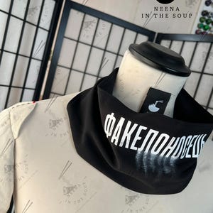 May include: A black neck gaiter with white Cyrillic text. The gaiter is draped on a mannequin. A small tag with a logo is visible. The background includes a black folding screen and a logo.
