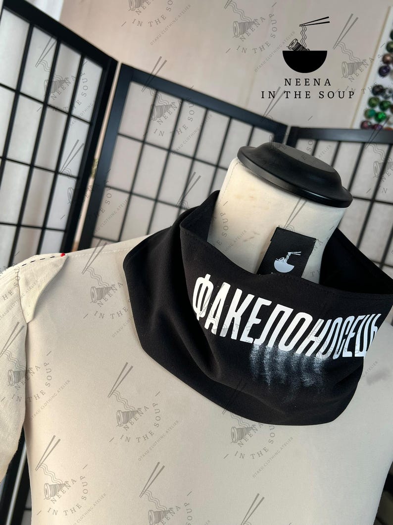 May include: A black neck gaiter with white Cyrillic text. The gaiter is draped on a mannequin. A small tag with a logo is visible. The background includes a black folding screen and a logo.