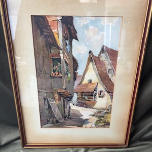 May include: A framed watercolor painting depicting a narrow street scene with timber-framed buildings, brown roofs, and a light blue sky. The artwork features a window with red flowers and is enclosed in a thin, dark red wooden frame with gold trim.