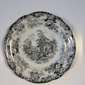 May include: A round ceramic plate featuring a black and white transferware pattern. The center depicts a landscape with a house and trees, while the scalloped rim is decorated with an intricate floral design of roses and leaves.