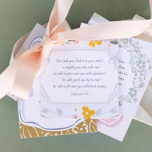 May include: A collection of printed cards tied with a light pink ribbon. The cards feature floral designs and text from Zephaniah 3:17. A small, off-white fabric pouch is also included. The background is a muted green.