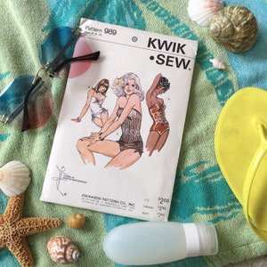 May include: A vintage sewing pattern for a swimsuit, featuring three women in different styles. The pattern is titled "Kwik Sew" and is for sizes 6-8-10. The pattern is on a white background with a blue and green beach towel, seashells, and sunglasses.