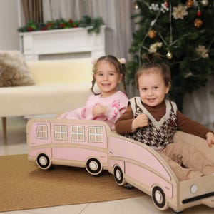 May include: Two children smiling in a pink wooden bus-shaped toy. The toy has black wheels and window cutouts. The children are wearing casual clothes. The background includes a couch, a Christmas tree, and a rug.