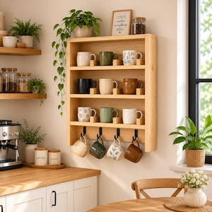 May include: A light-colored wooden wall-mounted mug rack featuring four horizontal shelves and four black metal hooks at the bottom. The rack holds twelve ceramic mugs in various colors and patterns, with a framed sign reading COFFEE makes everything better.