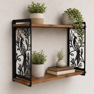Puede incluir: Una estantería de pared de dos niveles con dos tablas de madera horizontales sostenidas por paneles laterales de metal negro. Cada panel muestra un diseño cortado con láser de un pájaro sobre una rama. Plantas en macetas y dos libros descansan en los estantes.