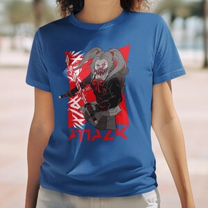 May include: A royal blue t-shirt with a graphic of a girl with pigtails wearing a black hooded jacket and holding a sword. The girl has a red and white mask on her face. The graphic is set against a red background with white jagged lines and the word 'ATTACK' written in white letters below the graphic.